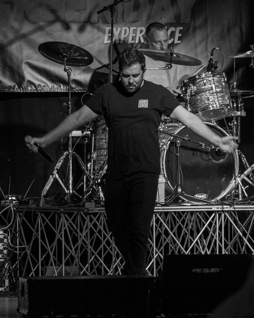 Black-and-white photograph of a vocalist with arms outstretched in front of a drum kit